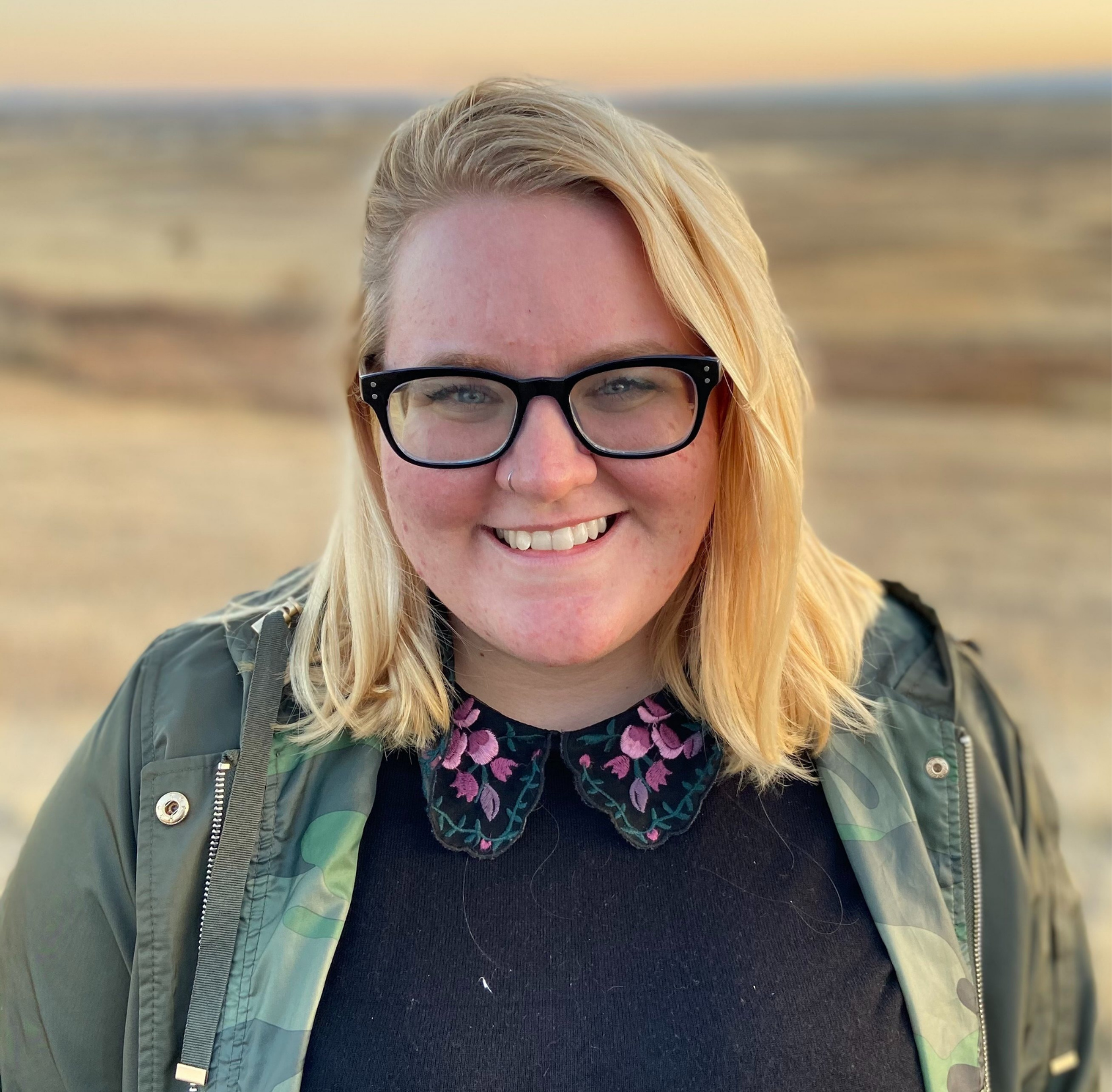 Zoe Henderson, a white woman with shoulder-length blonde hair and blue eyes. She is standing in a field and in front of the setting sun.